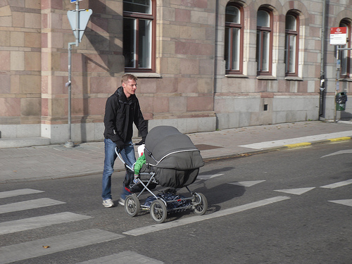 A thin blond man in jeans and a black jacket pushes a gray baby carriage toward us across a crosswalk in an otherwise empty street.