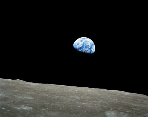 The Earth is seen as a small blue dot against a wide black sky, with the surface of the Moon in the foreground