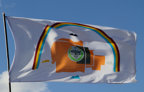 A white flag flies against a blue sky. The flag bears a map of the Navajo reservation, surrounded by four mountains, and a rainbow.